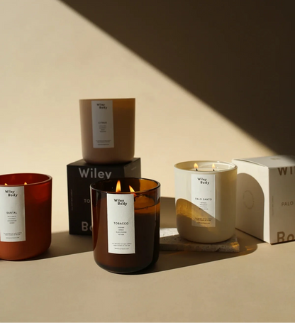 Wiley Body – Grupowe zdjęcie kolekcji produktów Tobacco: zapalona i niezapalona świeca sojowa w bursztynowych słoikach oraz kilka zamkniętych kartoników zapachowych z logiem marki w nastrojowym, dramatycznym świetle.