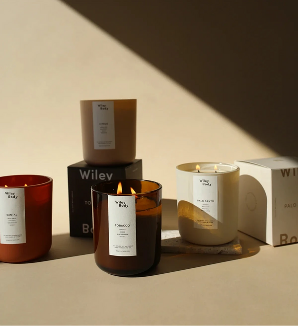 Wiley Body – Grupowe zdjęcie kolekcji produktów Tobacco: zapalona i niezapalona świeca sojowa w bursztynowych słoikach oraz kilka zamkniętych kartoników zapachowych z logiem marki w nastrojowym, dramatycznym świetle.